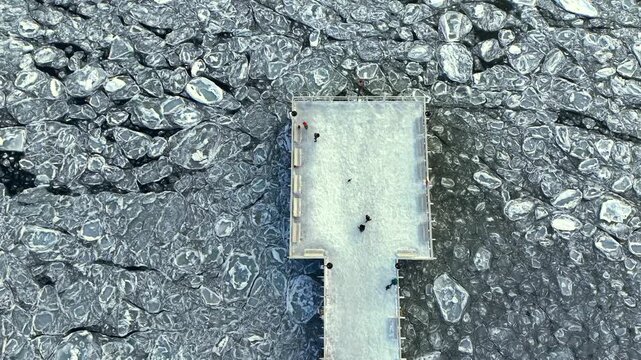 Aerial top-down drone shot of the Orłowo pier and walking people amid drifting ice floes on the frozen Baltic Sea in winter, Pomerania, Poland