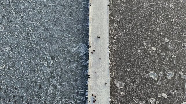Aerial top-down drone shot of the Orłowo pier as a line through abstract cracked ice on a frozen winter sea with people walking, Gdynia, Poland