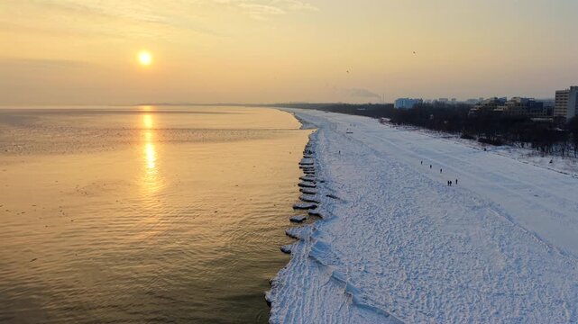 Aerial drone shot flying along the snow-covered Sopot beach toward the setting sun over the Baltic Sea at golden hour in winter, Poland