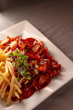 Close-up of white square plate holding french fries, beef pieces in red sauce, sliced peppers and microgreens on gray tabletop.