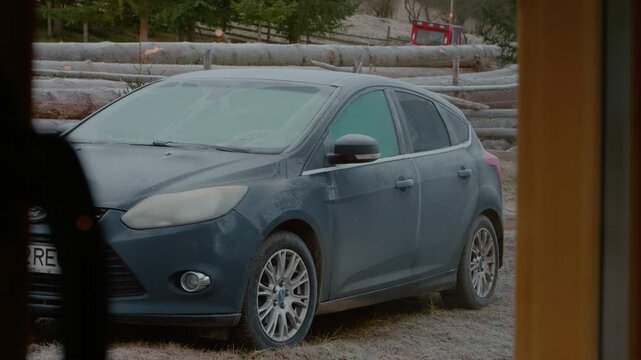 Frost forms on a parked car in the mountains early in the morning. Trees surround the area, and a wooden fence is visible in the background.