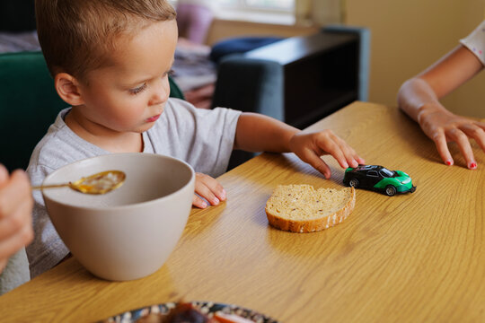 Cute toddler boy playing with a toy car while sitting at a kitchen table with a bowl of soup and bread during mealtime at home.