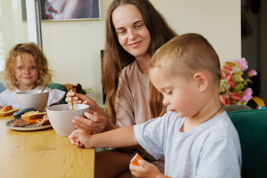 Caring mother holding a soup bowl while feeding her little son at a dining table during family breakfast in a bright kitchen.