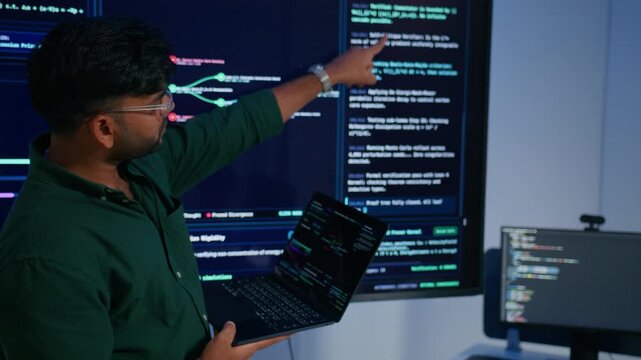 Male DevOps Engineer Holding Digital Tablet and Explaining Software Deployment Architecture and Discussing Development Operations Strategy with Colleagues Reviewing Programming Code on Large Screen.