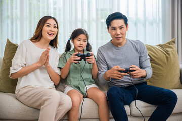 Happy Asian family enjoying a video game together on their couch, father mother and daughter sitting on a sofa playing games with joystick controller at home and having fun in living room © sorapop