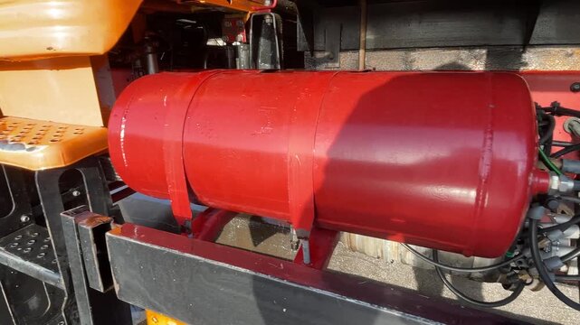 A red tank and its connected hoses are exposed along a truck chassis, showing the mounting hardware and pipe connections beside a black frame rail, highlighting truck washing in a wash yard.