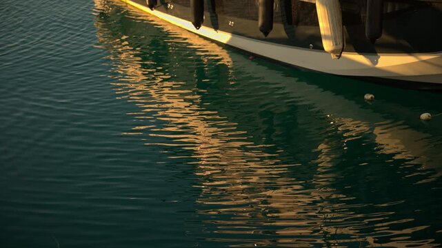 Golden sunlight reflecting on rippled water alongside the hull and fenders of a boat moored at a harbor during sunset. 4K.
