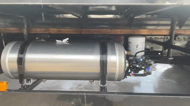 Water sprays beneath a cargo truck around a metal tank and chassis components, with the washing lance reaching into the space below the bed, documenting truck cleaning during routine upkeep.