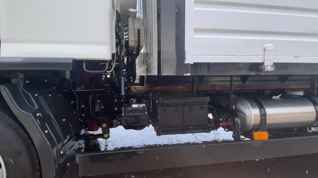 The space beneath a white truck cab reveals chassis rails, hoses and mounted components beside the fuel tank during a vehicle washing session, illustrating vehicle upkeep for road transport.