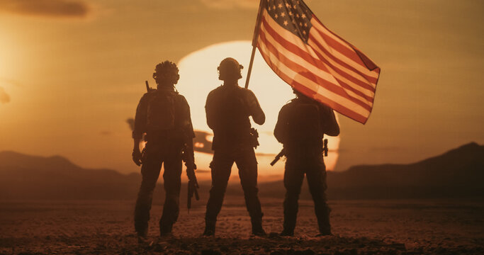 Backlit Infantry Unit of Three Armed Soldiers Pauses on Barren Desert Terrain, One Hoisting the American Flag on a Pole While Comrades Hold Rifles, Facing a Huge Burning Sun Over Distant Hills.
