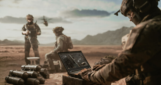 Gloved Soldier in Helmet Studies Live Aerial Surveillance Imagery on a Field Laptop Set Atop an Ammunition Crate, Mortar Rounds Piled Nearby as a Guard Watches and a Helicopter Hovers Over Desert.
