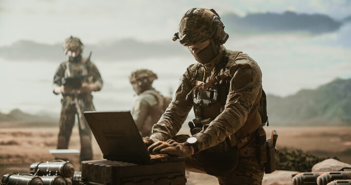 Masked Operator in Camouflage and Helmet Types Coordinates on a Rugged Laptop Resting on an Ammunition Crate at a Dusty Desert Outpost, Teammates Holding Rifles on Perimeter Security Behind.