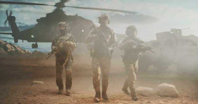 Three Armed Special Forces Soldiers in Camouflage and Helmets Advance on Foot Through Swirling Dust After Helicopter Insertion, Rifles Ready, With an Armored Vehicle Securing the Desert Zone.