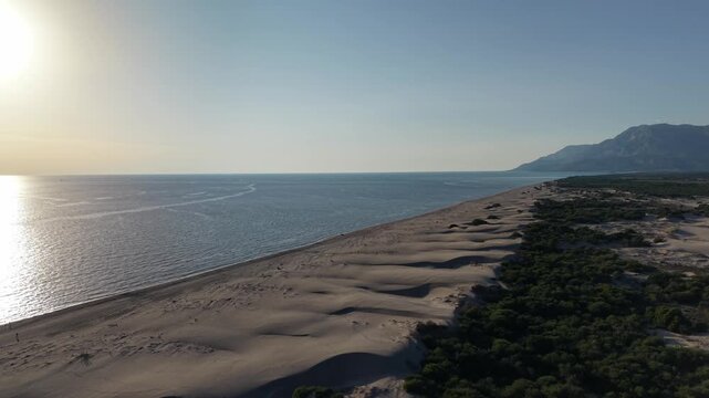 Aerial seaside scene showing long sandy dune ridge meeting calm ocean under late afternoon sun. Smooth wind-sculpted sand dunes run parallel to shoreline with scattered footprints and sparse driftwood