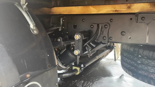 The area behind a truck fender reveals suspension brackets, bolts and chassis parts, showing water on the components during vehicle cleaning, illustrating vehicle washing for road transport.