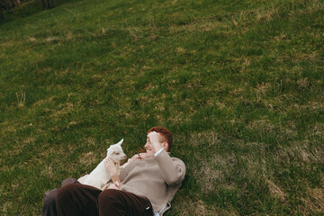 Fototapeta premium Cheerful man relaxing outdoors with a curious goat kid beside him, sunlit grass, casual clothing, warm smile, peaceful moment, nature setting, candid lifestyle scene