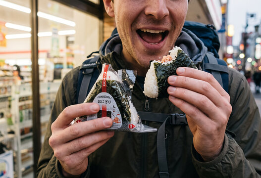 A foreign tourist happily tucking into a convenience store rice ball(onigiri). Their expression conveys the sheer delight of tasting it for the very first time since arriving in Japan.