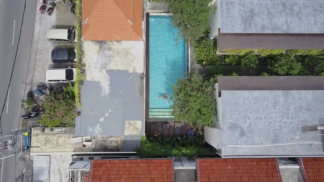 Rooftops, a street and a villa pool in Bali seen straight down as the camera descends to a woman floating on her back in the water, Aerial View