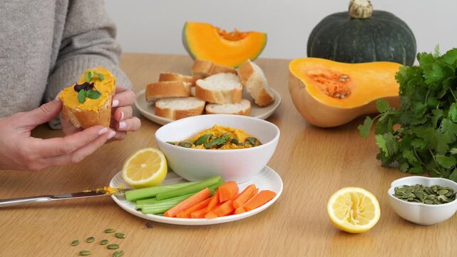 Hands spreading creamy squash dip on crusty bread with fresh vegetables and pumpkin seeds on wooden table, cozy autumn kitchen snack scene, closeup composition, natural light