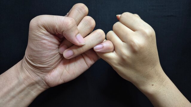 Close-up photograph of two human hands linking their little fingers in a pinky promise gesture against a black background. 