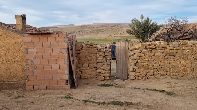 A traditional rural Algerian farm featuring rustic stone walls, mud-brick buildings, and a wooden gate leading to the house entrance under a cloudy sky.