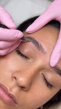 Close up of a beautician hands in pink gloves shaping eyebrows of a woman with a permanent makeup pen tool, cosmetic procedure, beauty concept.
