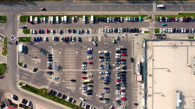 Overhead aerial drone shot showing cars moving and parking in a large, busy outdoor lot next to a shopping center, with traffic flowing on the adjacent city street and road infrastructure