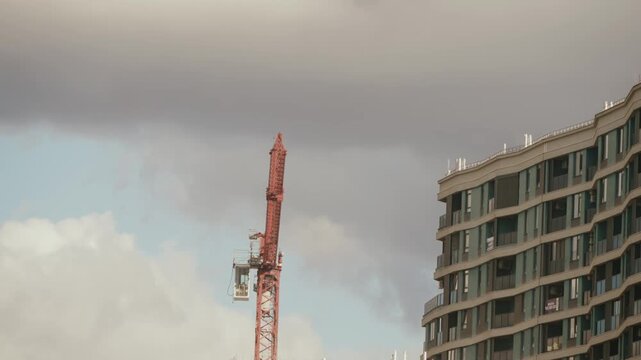 Red industrial tower crane slowly rotating next to a modern residential building with a curved facade, under a cloudy sky, symbolizing urban development and construction progress. timelapse