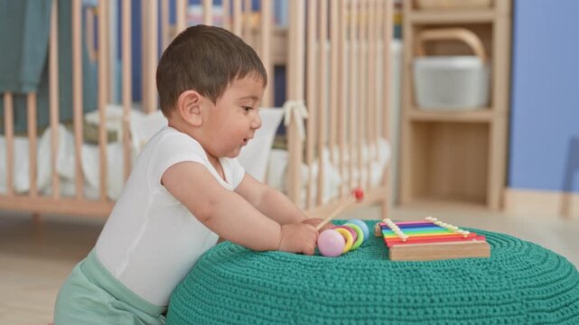 Hispanic toddler boy leaning on teal pouf, playing with a wooden xylophone and ring stacker in a building nursery; curiosity.