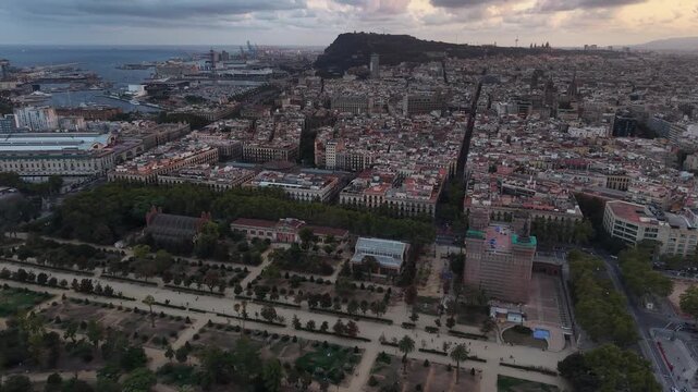 Barcelona cityscape sprawls from tree-lined Parc de la Ciutadella gardens toward dense historic rooftops, a busy harbor, and Montjuic hill beneath a cloudy evening sky. Aerial drone approaching shot