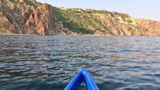 Rugged coastal cliffs rise above calm Black Sea waters as a blue kayak prow glides forward. Forward-facing point-of-view tracking shot.