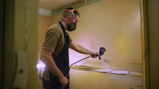 Man paints wall with spray gun, wearing mask and overalls. Light shines on his focused, steady motion. Room shows unfinished walls, bright work area. Great for construction, renovation