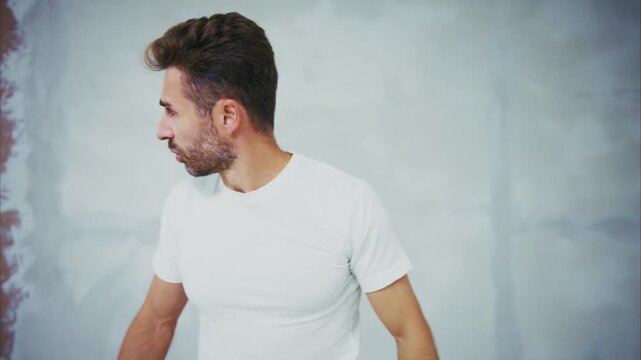 Man paints wall with brush, focused on task. He wears white shirt and pants, shows concentration. Wall is partially covered in light gray paint. Background shows unfinished room with red paint streaks