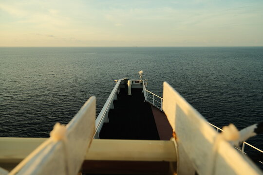 View over a ship's bow toward the open sea in warm evening light, with deck railings and onboard equipment creating depth and perspective in a maritime setting.