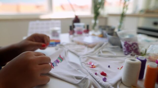 Closeup of hands placing colorful beads onto white garment on sunlit kitchen table, showing DIY embellishment on fabric with careful bead-by-bead arrangement