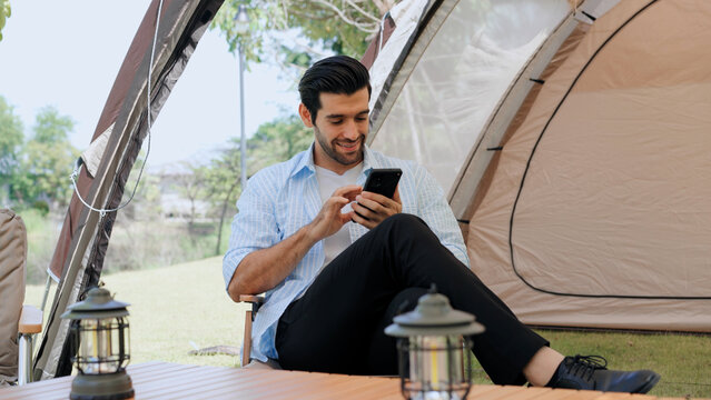 Happy young man using smartphone while relaxing outside his camping tent in nature, enjoying mobile connectivity, digital nomad lifestyle, outdoor recreation, and peaceful vacation time