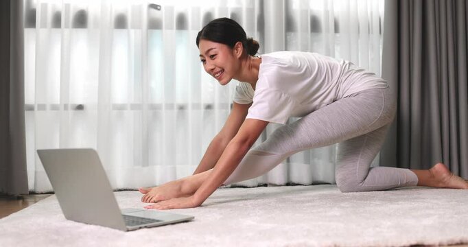 Asian woman practicing online yoga and stretching exercises watching a laptop lesson on a carpet at home, promoting healthy lifestyle, fitness training, flexibility, wellness, and self-care routine