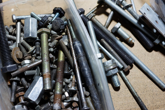 Assorted metal hardware including various bolts, nuts, threaded rods, and fasteners scattered inside a plastic storage container and across a workshop surface for home improvement projects