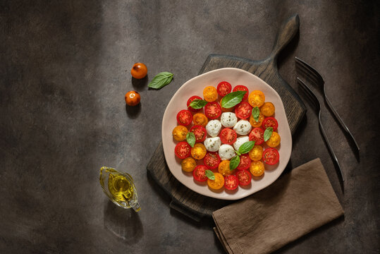 Caprese salad with mozzarella, cherry tomatoes and basil leaves in a plate on a dark grunge background with sunlight, top view