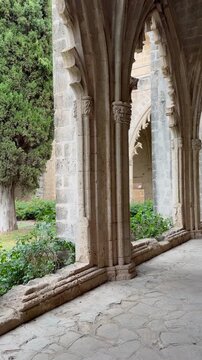 Kyrenia, Cyprus - October 3, 2026 Bellapais Abbey cloister frames quiet garden courtyard through pointed stone arches. Camera movement reveals cypress trees, greenery and weathered medieval masonry.