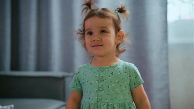 Curly-haired toddler stands near couch, smiling. She wears a green dress with small patterns. Her pigtails bounce as she moves toward toys. Soft background shows curtains and cushions