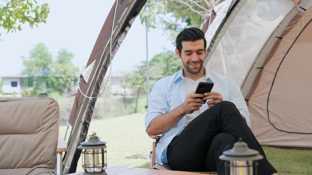 Happy young man using smartphone while relaxing outside his camping tent in nature, enjoying mobile connectivity, digital nomad lifestyle, outdoor recreation, and peaceful vacation time