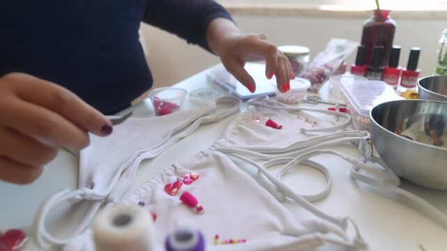 Close-up hands placing pink purple beads and sequins onto white garment, securing with small metal tool on tabletop craft workspace with thread and nail polish nearby