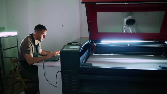 Focused worker operates laser cutter, monitoring process. Machine glows with precision light, showing active work. Man concentrates, hands on laptop, eyes on screen. Ideal for manufacturing