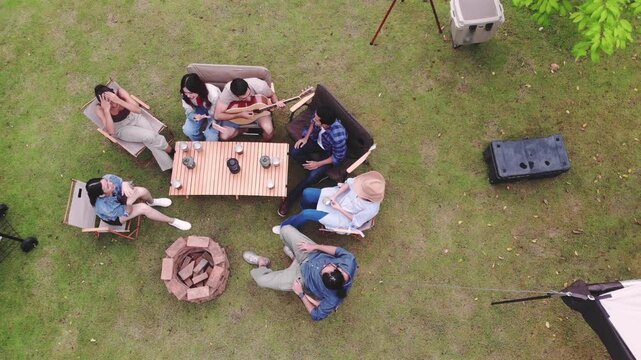 Aerial top view of diverse happy friends sitting around a table playing acoustic guitar, singing, and enjoying a outdoor camping party on green lawn during a weekend holiday vacation