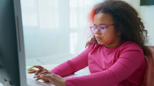 Young girl with glasses focuses on screen. She types with intent, hands steady. Soft light spills from window behind. Her pink shirt matches her cheerful tone. Perfect for education, remote learning