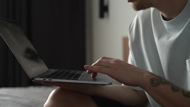 A man in a light T-shirt works on a laptop while sitting comfortably in a bright room. The scene represents remote work, freelancing, online business, and the everyday lifestyle of IT professionals