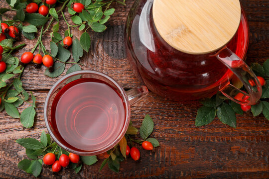 Aromatic rosehip tea in glass cup, teapot, berries and green leaves on wooden table, flat lay
