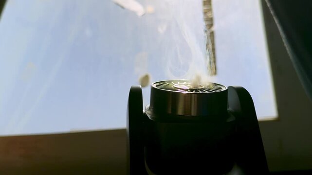 Cinematic close up of a black diffuser emitting white smoke indoors. The device forms a dark silhouette against a bright window illuminated by natural daylight.