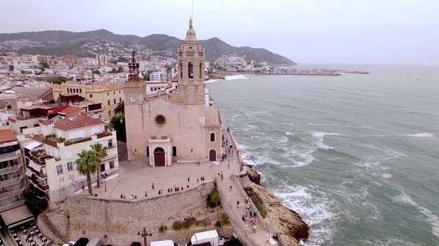 4K drone aerial view of Sitges, Spain, featuring the iconic Sant Bartomeu i Santa Tecla church overlooking the Mediterranean coast, beaches and historic seaside town.